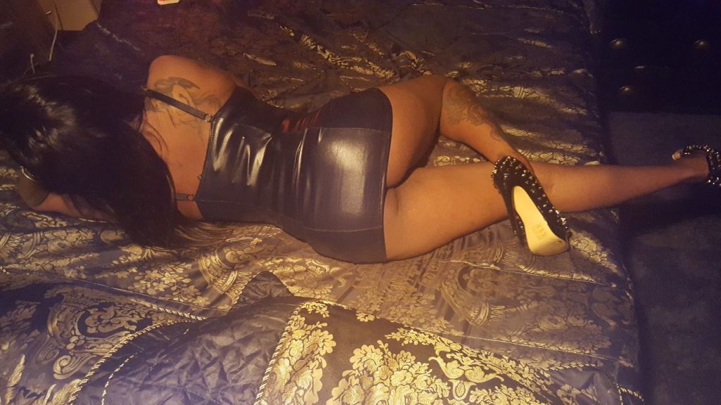Nicky on her bed in a black leather dress showing her